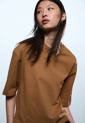 Mladá žena s dlhými tmavými vlasmi, ktorá má na sebe hnedú voľnú krátkym rukávom košeľu, pozerá sa do strany na jednoduchom svetlom pozadí.