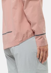Giacca rosa chiaro con una texture liscia, caratterizzata da una striscia di accento grigia all'orlo e alle maniche, abbinata a pantaloni corti grigi chiari.