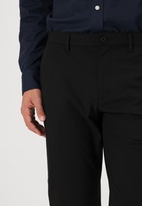 Černé kalhoty s hladkým povrchem, mají pružný pas a přední kapsy. Model má na sobě modrou vzorovanou košili.