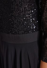 Robe noire avec un corsage en sequins, une taille noire lisse et une jupe plissée drapée, mettant en avant une texture scintillante et un design élégant.