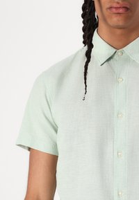 Camicia verde chiaro a maniche corte con bottoni, realizzata in un tessuto testurizzato, dotata di colletto classico e dettagli di cucitura sottili.