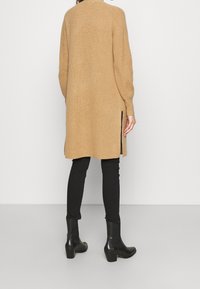 Lång, beige stickad kavaj med hög hals, sidoplen och ribbad textur. Matchas med svarta figurnära byxor och kängor med blockklack.