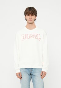 Mladý muž s kučeravými vlasmi v bielom pulóvri s červeným nápisom „DIESEL“ a svetlomodrých džínsoch, stojaci pred jednoliatym bielym pozadím.