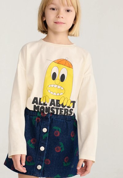 Enfant aux cheveux blonds courts portant un haut crème à manches longues avec un monstre jaune de dessin animé et une jupe bleue à motifs floraux rouges.