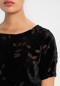 Haut en velours noir à manches courtes, présentant un motif de feuilles transparent, un col large et une texture douce.
