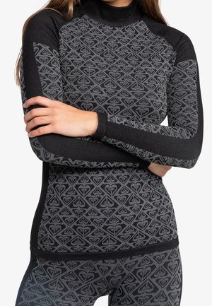 Frau trägt ein eng anliegendes schwarz-grau gemustertes Langarmshirt mit passenden Leggings, Arme vor der Brust verschränkt.