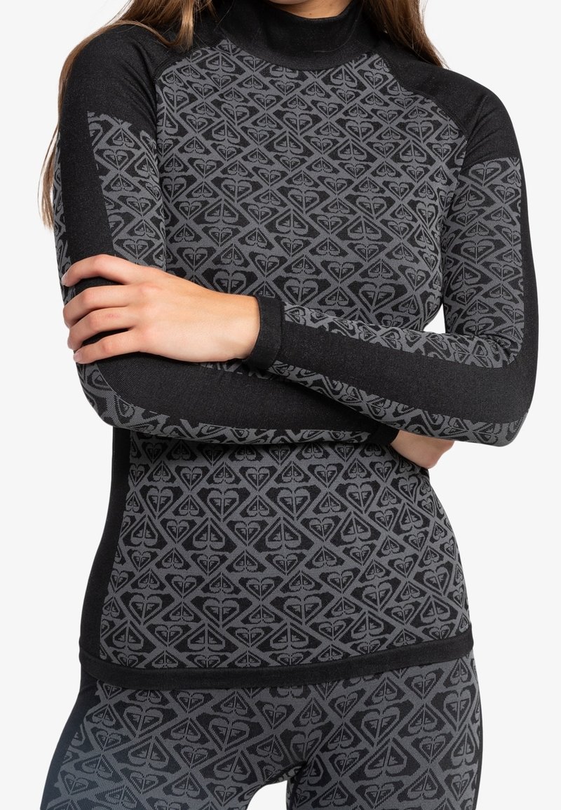 Femme portant un haut moulant à manches longues noir et gris à motifs avec des leggings assortis, bras croisés sur la poitrine.
