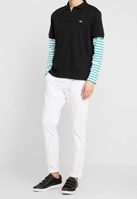 Polo shirt noir à manches courtes, superposé à une chemise à rayures turquoise et blanche à manches longues. Un pantalon blanc et des sneakers noires complètent le look.