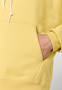 Felpa gialla con tasca a marsupio, cappuccio con lacci, polsini a costine e una texture morbida. Il tessuto appare liscio e leggero.