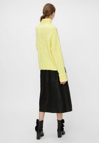 Pull en maille jaune avec un col haut et un détail texturé ; associé à une jupe midi en cuir noir et des bottines noires à volants.