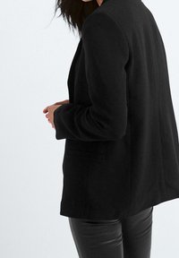 Blazer noir à coupe ajustée, avec une fente unique au dos, des épaules structurées et des poches latérales. Texture de tissu lisse avec une finition mate.