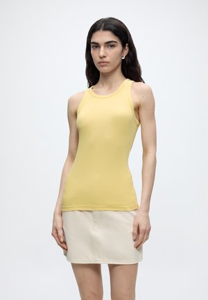 Femme aux longs cheveux foncés portant un haut jaune sans manches et une jupe beige clair, debout devant un fond blanc uni.