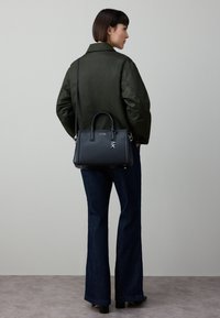 Svart skinnveske med to håndtak og avtakbar skulderrem, med sølvfarget metallbeslag. Modellen har på seg en mørkegrønn jakke og jeans.