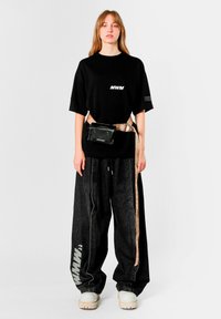 Camiseta negra de gran tamaño con logotipo blanco; pantalones vaqueros negros de pierna ancha; riñonera beige; zapatos blancos gruesos; estilo urbano casual.