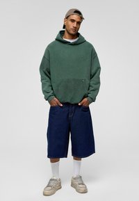 Groene hoodie met een buidelzak, gecombineerd met losse, donkere denimshorts. Witte enkelsokken en grijze sneakers maken de outfit compleet.