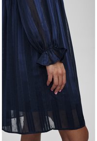 Granatowa sukienka z przezroczystej, paskowanej tkaniny. Posiada marszczone rękawy, rozkloszowane mankiety i wzorzysty materiał. Dłoń ułożona na tkaninie.