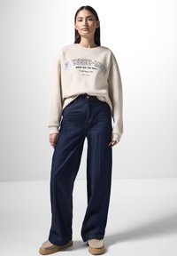 Felpa crema con testo grafico, abbinata a jeans scuri in denim a vita alta e gamba larga, e scarpe slip-on beige con foderatura testurizzata.