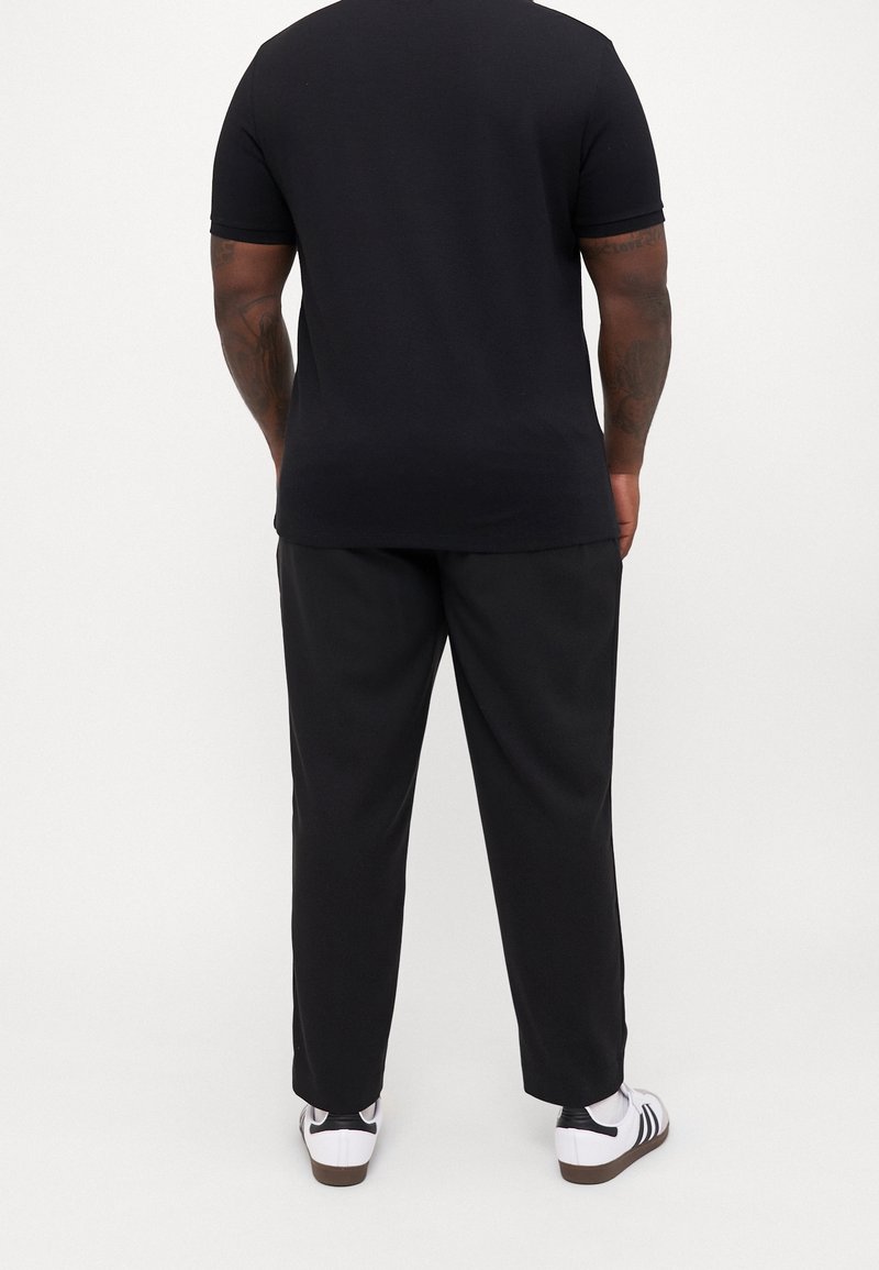 Homme debout de dos, portant une chemise noire à manches courtes, un pantalon noir, des chaussettes blanches et des baskets blanches à rayures noires.