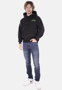 Felpa nera con tasca frontale, logo verde sul petto, abbinata a jeans blu e sneakers bianche. Modello in piedi davanti a uno sfondo neutro.