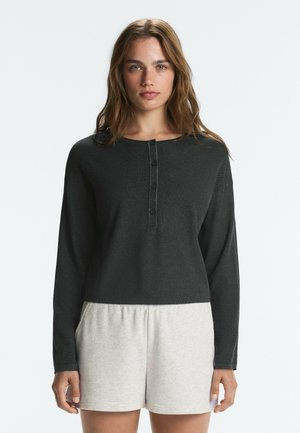 Jeune femme aux cheveux bruns ondulés portant une chemise noire à manches longues boutonnée et un short gris clair, debout devant un fond clair uni.