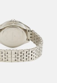 Sølv rustfrit stål ur med en rund kasse, tekstureret krone og et mesh-armbånd. Bagsiden har indgraverede branddetaljer og information om vandmodstand.