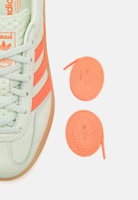 Minzgrüner Sneaker mit orangefarbenen Streifen, strukturiertem Zungenbereich und hellgrauen Schnürsenkeln. Zwei runde orangefarbene Schnürsenkel liegen neben dem Schuh.