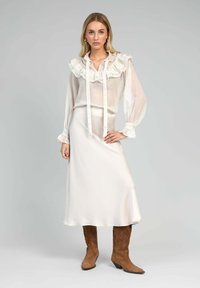 Blouse blanche transparente avec col à volants et détail de nœud, associée à une jupe midi crème lisse et des bottes style western marron.