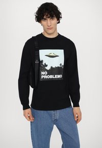 Black long-sleeve T-shirt featuring a graphic of a UFO over trees with “NO PROBLEMO” text. Paired with blue denim jeans and a black shoulder bag.
