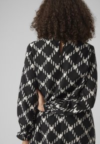 Melnā un baltā rakstainā kleita ar dimanta dizainu, kas ir ar garām piedurknēm, elastīgām aprocēm un atslēgas cauruma aizmugures detaļu.