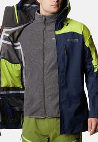 Giacca da uomo con un esterno blu navy e accenti verde brillante, chiusura con zip e fodera in felpa grigia chiara. Include tasche e cappuccio.