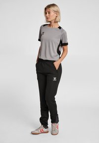 Graues Sportoberteil mit schwarzen Akzenten, kombiniert mit schwarzen Jogginghosen mit seitlichen Taschen. Helle Sneakers mit roten Details.