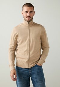 Bežinis zip-up megztinis su aukštu apykakle, ilgomis rankovėmis ir raštuotais rankogaliais, derinamas su mėlynomis džinsinėmis kelnėmis. Minkšta tekstūra ir prigludusi forma.