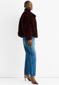 Woman wearing a dark burgundy faux fur jacket, blue straight-leg jeans, and matching burgundy high heel slingback shoes, standing sideways.