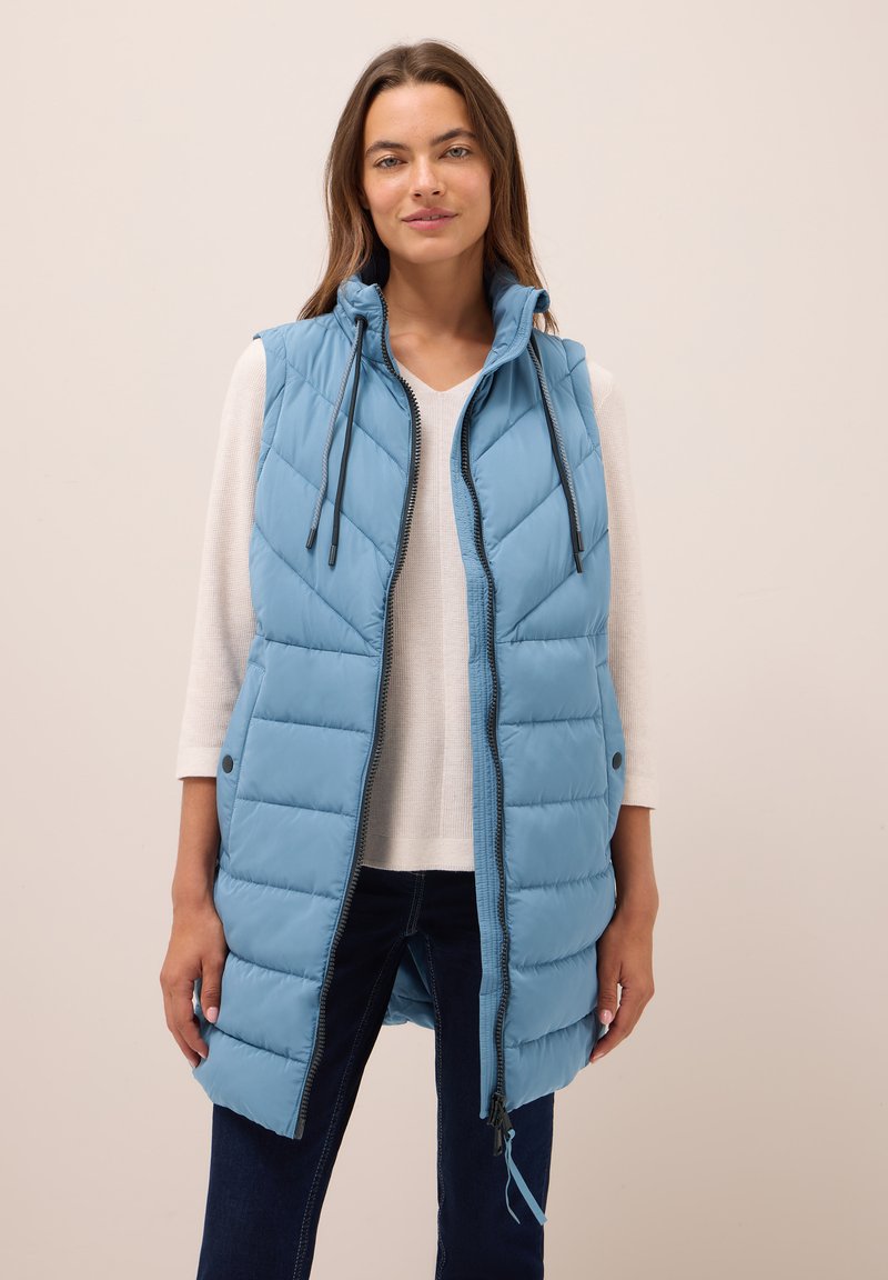 Gilet matelassé bleu clair avec un col montant, fermeture éclair et cordons de serrage. Doté d'une texture matelassée et de poches latérales, porté sur un haut blanc.