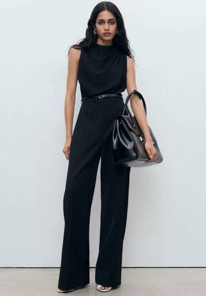 Sort jumpsuit med høj hals, ærmeløst design og vide ben, parret med en struktureret sort håndtaske. Glat stof med en tætsiddende talje.