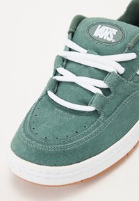 Baskets en daim vert avec des lacets blancs, bout rond et détails perforés. Présente un patch logo Vans sur la languette et une semelle en caoutchouc gaufré.