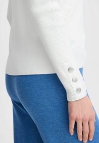 Un haut à manches longues blanc côtelé avec trois boutons argentés au niveau des poignets, associé à un pantalon bleu texturé. L'encolure est de style ras du cou.