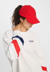 Vrouw met krullend haar in een rode pet en een witte Hummel-sweater met rode en marineblauwe strepen, die naar beneden kijkt en haar hals aanraakt.