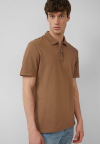 Braunes Polohemd mit kurzen Ärmeln und einem Reißverschlusskragen, aus strukturierter Baumwolle. Trägt man zu hellblauen Jeans.