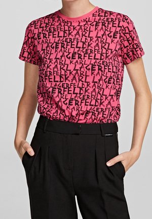 Osoba ubrana w różową koszulkę z krótkim rękawem z czarnym wzorem tekstowym „Karl Lagerfeld” oraz czarne spodnie z wysokim stanem, z rękami w kieszeniach.