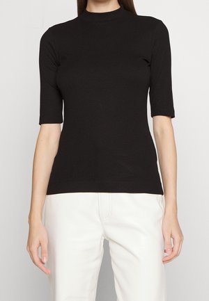 Femme portant un haut noir côtelé à manches courtes avec un col montant et un pantalon blanc, montrée des épaules aux hanches sur un fond uni.
