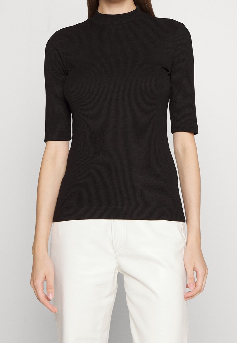 Femme portant un haut noir côtelé à manches courtes avec un col montant et un pantalon blanc, montrée des épaules aux hanches sur un fond uni.