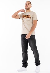 Kreminės spalvos medvilnės marškinėliai su rudu „Lonsdale London“ logotipu, derinami su tamsiai mėlynomis džinsais ir baltais sportbačiais. Modelis pozavo laisvai.
