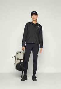 Kvinne som har på seg en svart North Face-jakke, tights, caps og turstøvler, og holder en lys grå ryggsekk mot en ensfarget bakgrunn.