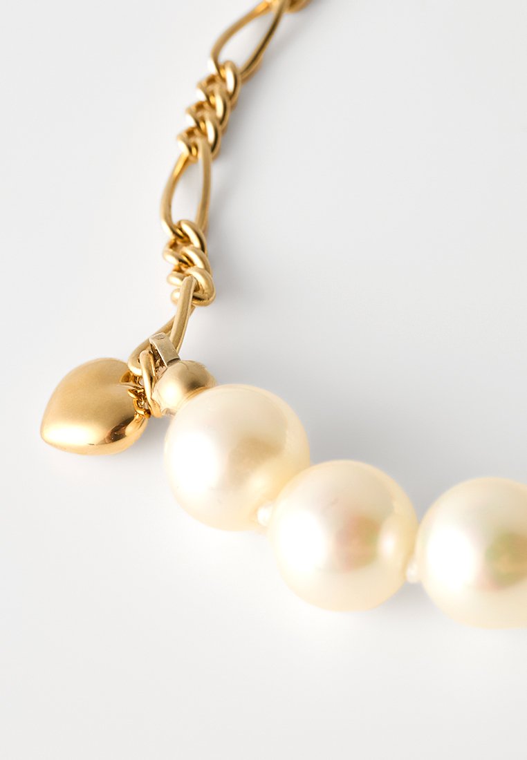 Gouden ketting met glanzende witte parels en een klein hartvormig gouden bedeltje, met een gestructureerd kettingontwerp.