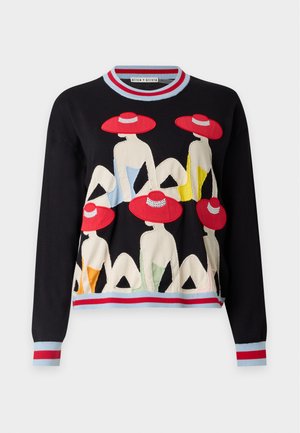 Femme portant un pull noir avec des motifs de chapeau rouges et un jean large bleu avec une grande broderie florale rouge, debout devant un fond uni.