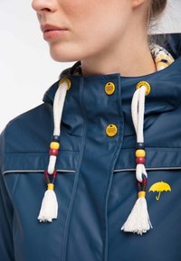 Lähikuva henkilöstä, joka käyttää tummansinistä sadetakki, jossa on valkoiset punotut narut,värikkäät helmet, keltaiset napit ja keltainen sateenvarjon logo.