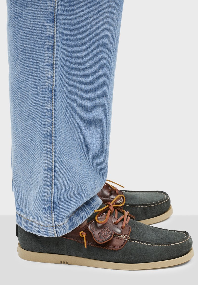 Zapatos de hombre en ante gris oscuro con detalles en piel marrón, suela beige y costura decorativa. Combinados con jeans de color azul claro.