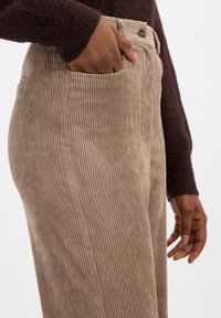 Pantalons en velours côtelé marron avec des côtes verticales, dotés d'une poche latérale et d'une fermeture à bouton à la taille, tenus par une main.