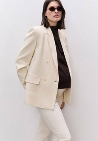 Krämfärgad dubbelknäppt blazer med slag, bärs över en mörkbrown topp, matchad med ljusa byxor. Modellen har på sig ovala solglasögon.